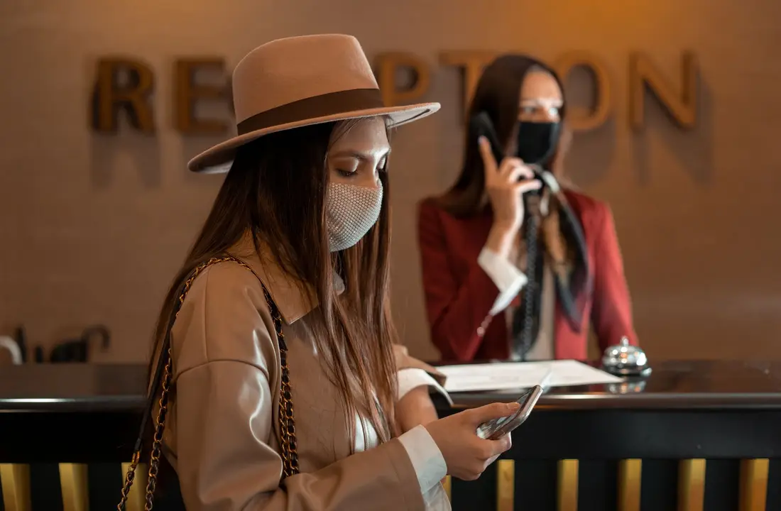 Woman wearing a mask checking her phone at a hotel reception during COVID-19.